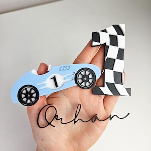 May include: A light blue race car and a number one cake topper with a black and white checkered design. The race car has black wheels and the number one has a checkered pattern. The name "Qurban" is written in cursive.