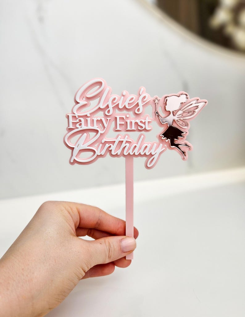 May include: A pink cake topper with the text "Elsie's Fairy First Birthday" in a layered design. A fairy silhouette with wings is on the right. The topper is held by a pink stick.
