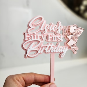 May include: A pink and white layered cake topper with the text "Elsie's Fairy First Birthday" and a fairy illustration. The topper is held by a pink stick.
