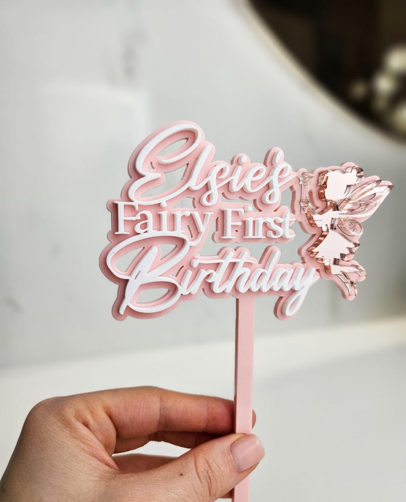 May include: A pink and white layered cake topper with the text "Elsie's Fairy First Birthday" and a fairy illustration. The topper is held by a pink stick.
