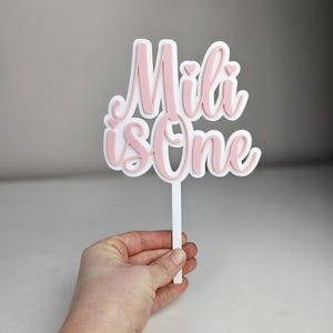 以下が含まれることがあります： ピンクと白のケーキトッパーで、「Mili is one」という文字が筆記体で書かれています。トッパーは白い棒に付いています。