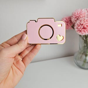 May include: A pink camera-shaped keychain with a gold outline and a small gold heart on the side.