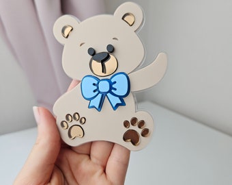 May include: A beige teddy bear with a blue bow tie and gold paw prints. The bear is made of acrylic and has a whimsical, cartoonish style.