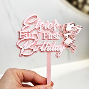 May include: A pink and white cake topper with the text "Elsie's Fairy First Birthday". A fairy with rose gold wings and a wand is on the right side. The topper is held by a pink stick.