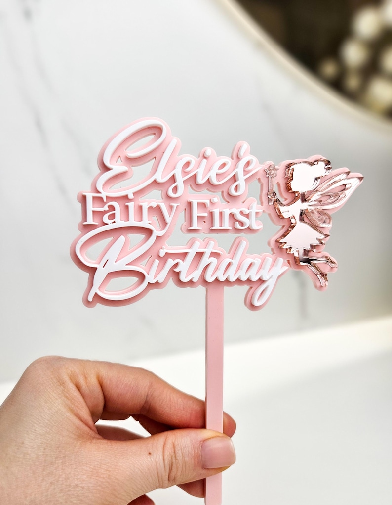 May include: A pink and white cake topper with the text "Elsie's Fairy First Birthday". A fairy with rose gold wings and a wand is on the right side. The topper is held by a pink stick.