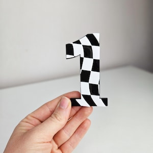 May include: A black and white chequered number one. The number is held in a hand against a white background. The number has a racing flag design.