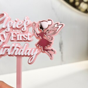 May include: A pink and white cake topper with the text "Elsie's First Birthday" and a fairy illustration. The fairy is in a pink dress with silver wings and a wand. The topper is designed for a birthday cake.