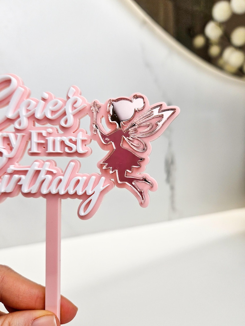 May include: A pink and white cake topper with the text "Elsie's First Birthday" and a fairy illustration. The fairy is in a pink dress with silver wings and a wand. The topper is designed for a birthday cake.