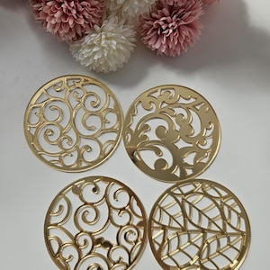 May include: Four gold-coloured, circular coasters with intricate, openwork designs. Each coaster features a unique pattern of swirling lines and geometric shapes. The coasters are displayed on a white surface, with pink and white flowers in the background.