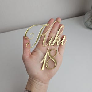 以下が含まれることがあります： 「Kiki」という名前と「18」という数字が書かれた、金色のアクリル製のネームプレート。筆記体で書かれています。