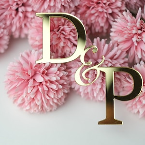 May include: Gold D & P wedding cake topper with a decorative script ampersand, surrounded by pink silk flowers.