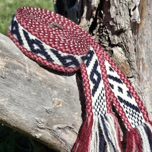 May include: A woven belt with a geometric pattern in burgundy, navy, and white. The belt is rolled up, displaying the intricate design and fringed ends. The belt is made of a thick, textured material.