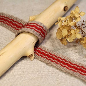 Puede incluir: Primer plano de una cinta tejida roja y beige atada alrededor de una rama de madera. La cinta es texturizada y tiene un brillo sutil. La rama es de color marrón claro y tiene un aspecto natural y rústico.