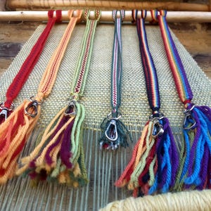 Peut inclure: Six lanières tissées colorées avec des glands et des fermoirs métalliques suspendus à un métier à tisser en bois. Les lanières sont tissées avec des fils de différentes couleurs, notamment le rouge, l'orange, le jaune, le vert, le bleu et le violet.