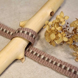 Puede incluir: Primer plano de una cinta tejida marrón y blanca atada alrededor de un trozo de madera a la deriva. La cinta tiene un sutil patrón de rayas marrones y blancas. Hay flores secas en el fondo.