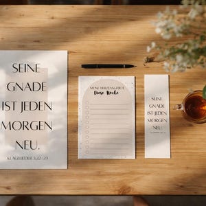 Puede incluir: Un juego de papelería con un tema motivacional, que presenta una impresión grande, un bloc de notas y un marcador, todos con el texto "SEINE GNADE IST JEDEN MORGEN NEU." Un bolígrafo y una taza de té también están sobre la superficie de madera.