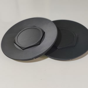 Puede incluir: Dos objetos negros y circulares con un centro hexagonal. Los objetos tienen una forma plana, similar a un disco, con una característica hexagonal en relieve en el centro. Probablemente son componentes o piezas de un mecanismo más grande.