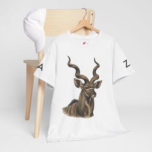 May include: A white t-shirt featuring a detailed illustration of a kudu with large spiraled horns on the chest and the letter A on the left sleeve and Z on the right sleeve, accompanied by a plain white baseball cap hanging on a wooden chair.