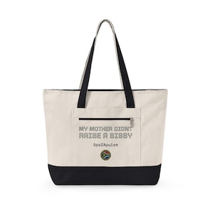 Pode incluir: Uma bolsa tote de lona na cor creme com alças e base pretas. A parte frontal apresenta um bolso com zíper com o texto "MY MOTHER DIDNT RAISE A SISSY" em preto, seguido por "SpaZApulse" e um pequeno emblema circular da bandeira da África do Sul.