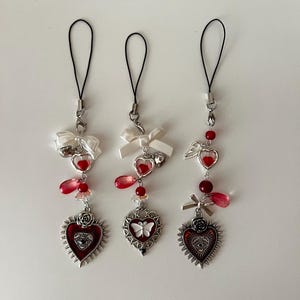 May include: Three decorative charms with black loops, white bows, and red heart-shaped pendants. Each charm features a different heart design, including a rose, a butterfly, and an eye. The charms are adorned with red beads and teardrop-shaped accents.