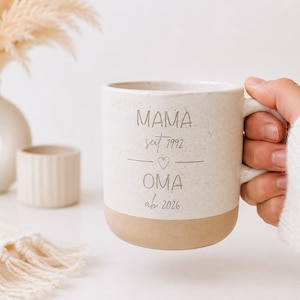 Könnte beinhalten: Eine gesprenkelte Keramiktasse mit beigem Boden, gehalten von einer Person in einem weißen Pullover. Auf der Tasse stehen die Worte "MAMA seit 1992 OMA ab 2026". Im Hintergrund befinden sich eine Vase und ein kleiner Keramiktopf.
