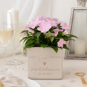 Könnte beinhalten: Ein quadratischer, hellfarbener Pflanzkasten mit rosa Blüten und grünen Blättern. Auf der Box stehen die Worte "LASST LIEBE WACHSEN" und die Namen "Josi & Johannes" mit dem Datum "04. JULI 2025" eingraviert. Die Szene umfasst ein Weinglas und eine Kerze.