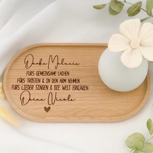 Könnte beinhalten: Ein ovales Holztablett mit graviertem deutschem Text und einer kleinen Vase mit einer weißen Blume. Das Tablett trägt die Worte "Danke Melanie". Eine kleine, weiße, kugelförmige Vase hält eine weiße Blume.
