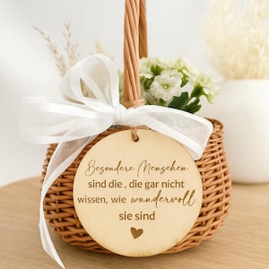 Könnte beinhalten: Ein Weidenkorb mit Henkel, gefüllt mit weißen Blumen und Grün, gebunden mit einem weißen Band. Ein Holzanhänger mit deutschem Text und einem Herz hängt am Korb.