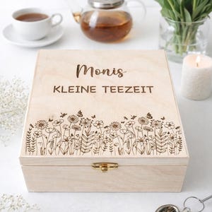 Könnte beinhalten: Eine hellfarbene Teebox aus Holz mit den Worten "Monis KLEINE TEEZEIT" auf dem Deckel eingraviert. Der Deckel hat ein Blumendesign. Eine Tasse Tee, eine Teekanne und ein Strauß weißer Blumen sind im Hintergrund.