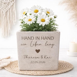Könnte beinhalten: Ein quadratischer, hellfarbener Topf mit Gänseblümchen. Der Topf trägt den Text "HAND IN HAND ein Leben lang" mit einem Herz, Namen und einem Datum. Der Topf steht auf einer gewebten Matte.
