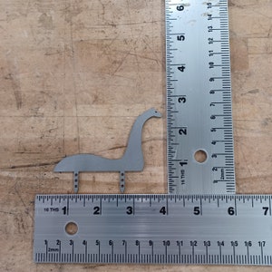 Op de afbeelding: Een metalen uitsnijding van een grijs Loch Ness-monster, ongeveer 7,5 cm hoog, met drie kleine poten. De uitsnijding is geplaatst op een houten oppervlak naast een liniaal.