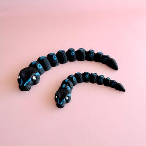 May include: Two black snake sculptures with blue detailing, set against a pink backdrop. The snakes feature segmented bodies and detailed heads, with blue markings around the eyes and along the spine. The sculptures are artistic and decorative.