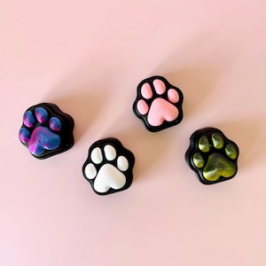 May include: Four paw-shaped decorative items with a black border and colourful paw prints. The paw prints are in shades of pink, white, blue, and green. The items are arranged on a light pink background.