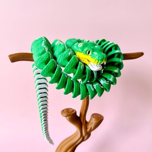 May include: A green and white articulated snake toy with yellow accents, coiled around a brown branch. The snake's head is detailed with yellow and white, set against a pink background. A fun, decorative item.