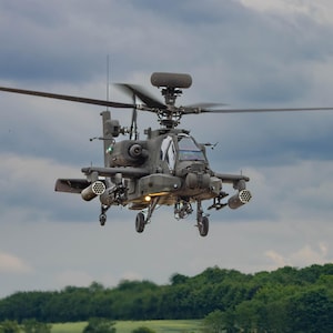 Könnte beinhalten: Ein dunkelolivgrüner militärischer Kampfhubschrauber im Flug vor einem bewölkten Himmel. Das Fluggerät verfügt über ein oben montiertes Radardom, einen Hauptrotor mit mehreren Blättern und seitlich angebrachte Raketenwerfer. Das Fahrwerk ist ausgefahren.