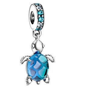 Può includere: Un charm in argento con un design a tartaruga marina. La tartaruga ha un guscio blu e turchese, e il charm è impreziosito da pietre preziose blu e trasparenti. Il charm è progettato per essere appeso a un braccialetto o una collana.