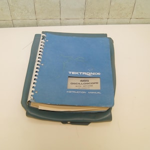 Könnte beinhalten: Ein blaues Tektronix 465 Oszilloskop-Handbuch mit Spiralbindung. Das Handbuch befindet sich in einem blauen Etui. Der Text auf dem Cover lautet "TEKTRONIX 465 OSCILLOSCOPE WITH OPTIONS INSTRUCTION MANUAL."