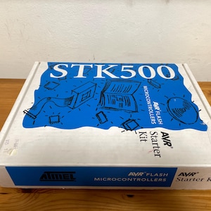 Peut inclure: Une boîte blanche avec du texte et des illustrations bleus et noirs. La boîte est étiquetée "STK500 AVR FLASH MICROCONTROLLERS Starter Kit".