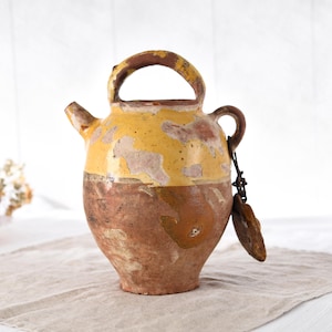 Peut inclure: Pichet en céramique antique et rustique, avec anse et bec verseur. Le pichet présente un design bicolore, avec une partie supérieure jaune et une partie inférieure marron. Une petite anse et un élément décoratif pendent sur le côté.