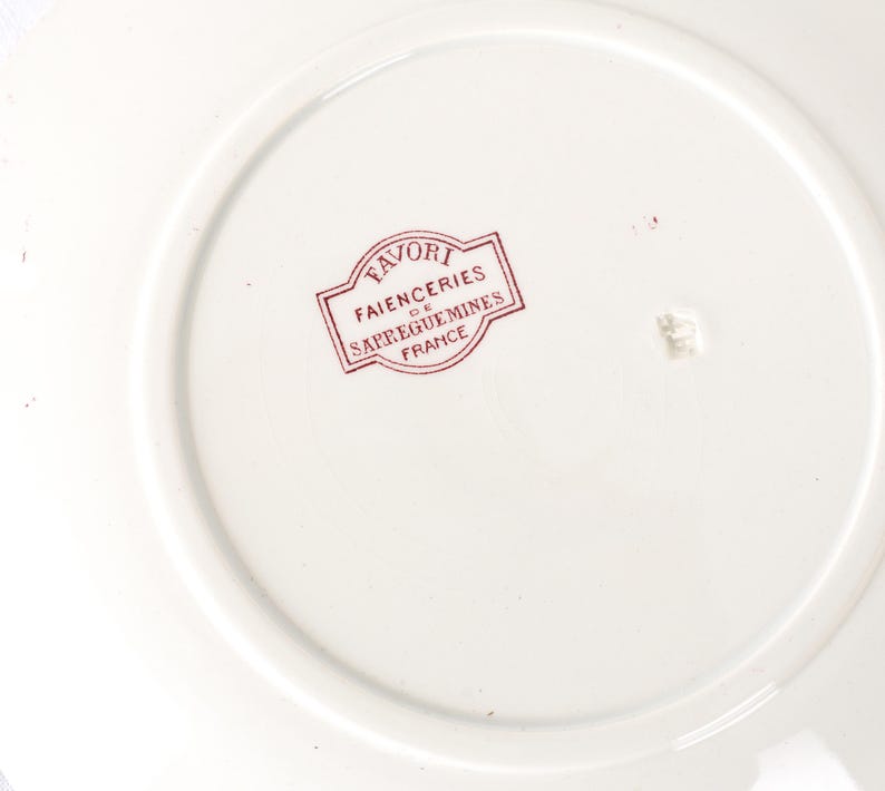 Puede incluir: Un plato de cer&aacute;mica blanco con un sello rojo que dice "FAVORI FAIENCERIES DE SARREGUEMINES FRANCE". El plato tiene un dise&ntilde;o circular con un borde elevado.