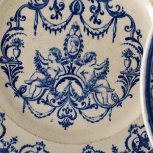 May include: Antique white and blue porcelain plate with a detailed design. The plate features a central motif of two cherubs, a portrait, and floral elements. The blue design is set against a white background, creating a classic aesthetic.
