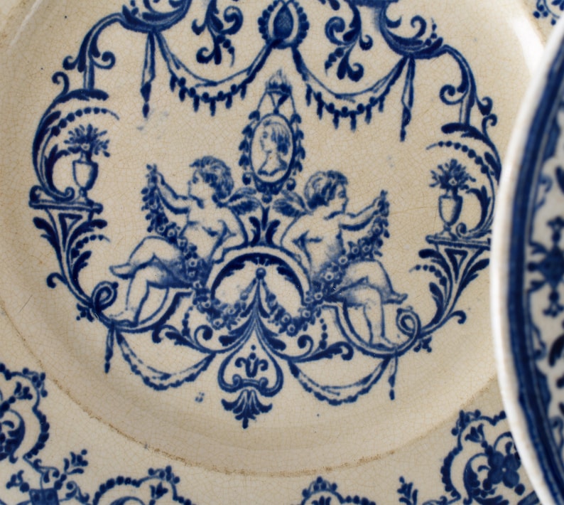 May include: Antique white and blue porcelain plate with a detailed design. The plate features a central motif of two cherubs, a portrait, and floral elements. The blue design is set against a white background, creating a classic aesthetic.