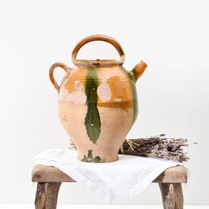 May include: A rustic, antique ceramic pitcher with a handle and spout. The pitcher features a color scheme of tan, green, and orange. It sits on a wooden stool with a white cloth and a bundle of dried lavender.