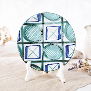 May include: A decorative ceramic plate with a geometric design. The plate features a grid pattern with squares and circles in shades of blue and teal, outlined in black. It is displayed on a white stand, showcasing its artistic design.