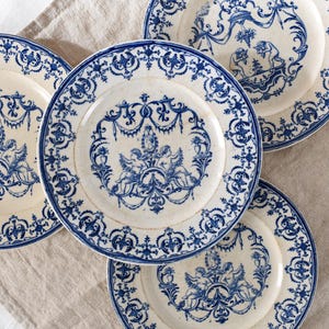 May include: A collection of antique white plates with blue decorative patterns. Each plate features a central design of cherubs and ornate scrollwork, surrounded by a detailed border. The plates are arranged on a neutral-colored linen surface.