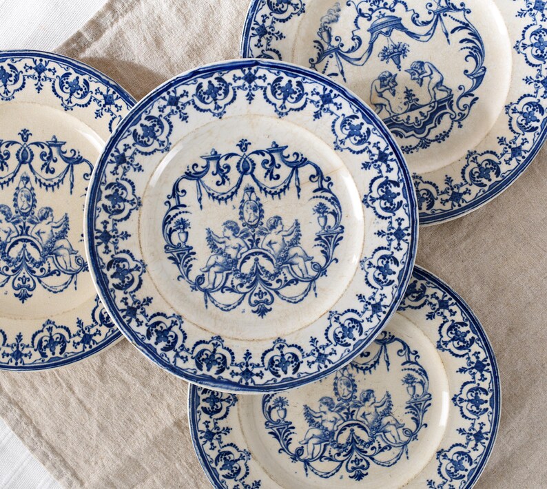 May include: A collection of antique white plates with blue decorative patterns. Each plate features a central design of cherubs and ornate scrollwork, surrounded by a detailed border. The plates are arranged on a neutral-colored linen surface.
