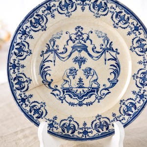 May include: Antique white and blue decorative plate with a detailed design. The plate features a central scene with two figures and ornate blue floral patterns around the rim. The plate is displayed on a white stand.