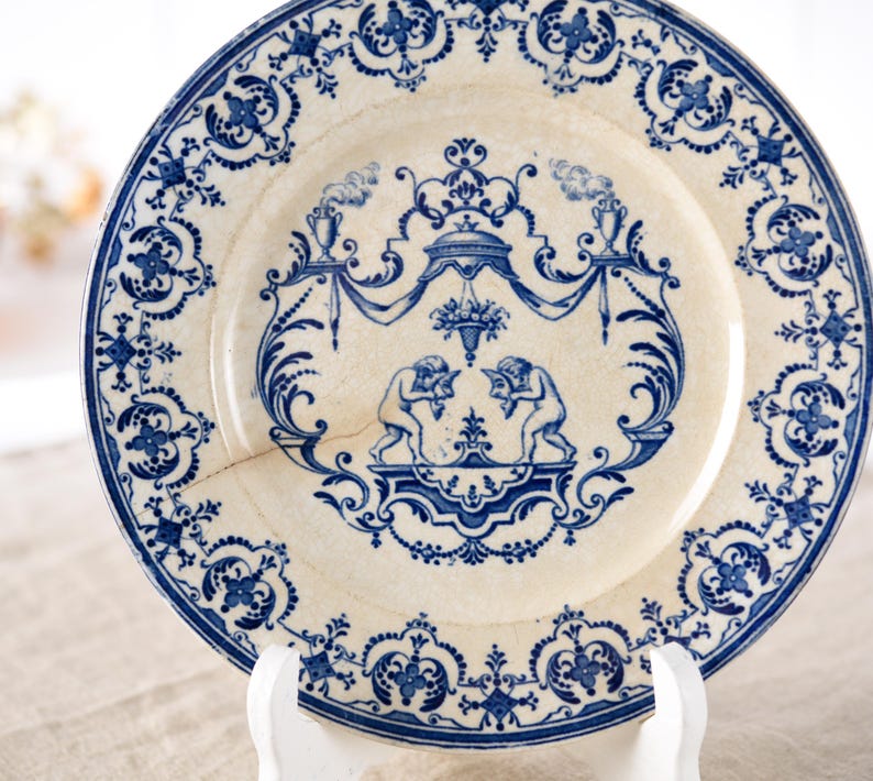 May include: Antique white and blue decorative plate with a detailed design. The plate features a central scene with two figures and ornate blue floral patterns around the rim. The plate is displayed on a white stand.