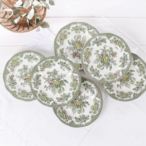 Pode incluir: Um conjunto de seis pratos de cerâmica branca com um desenho floral em verde, amarelo e azul. Cada prato tem uma borda ondulada e uma borda verde. Os pratos estão dispostos sobre um pano branco.