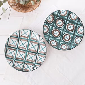 Puede incluir: Dos platos decorativos de cerámica con patrones geométricos. Uno presenta un diseño de cuadrícula turquesa y marrón con detalles blancos, mientras que el otro tiene un patrón de círculos turquesa y marrón. Los platos están sobre una superficie blanca.
